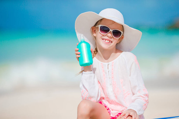 Pieni tyttö hattu päässä pullo aurinkovoidetta istuu solariumissa trooppisella rannalla
 - Valokuva, kuva