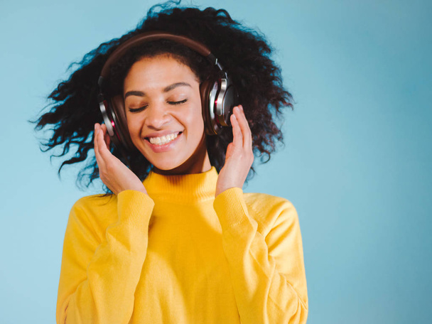 attractive african woman with headphones in dancing motion, hair fly, wearing yellow sweater, blue studio. Colorful picture, trendy girl - Fotó, kép