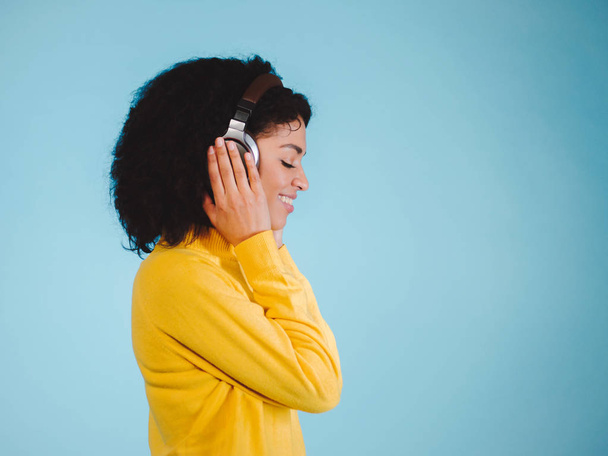 Beautiful young woman in headphones listening to music, smiling with closed eyes standing on a blue background in yellow sweatshirt - Foto, afbeelding
