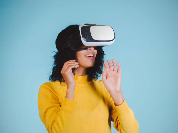 Future is right now. Confident young african american woman adjusting her virtual reality headset and smiling on blue background. Girl in VR goggles - Foto, imagen