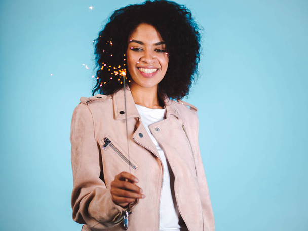 Sparkling Bengal fire in african american womans hand on blue background. Christmas Holiday Concept. Young pretty girl with afro hairstyle celebrating, smiling, enjoying time - Photo, Image