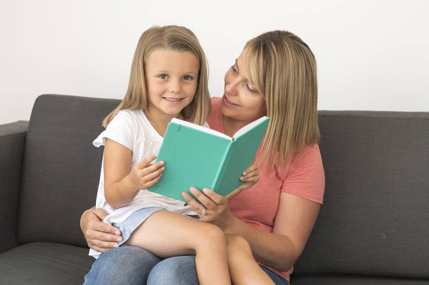 young beautiful and happy women sitting together with her adorable 7 years old adorable blond girl reading book enjoying telling story - Photo, Image