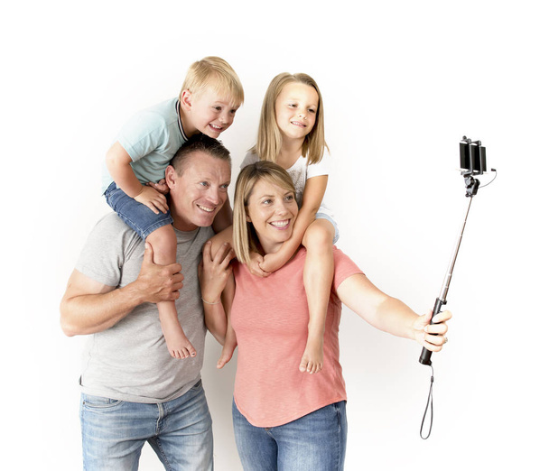 lovely young couple taking selfie photo self portrait with stick and mobile phone carrying son and daughter on shoulders posing happy smiling - Foto, Imagem