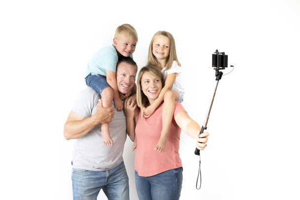 lovely young couple taking selfie photo self portrait with stick and mobile phone carrying son and daughter on shoulders posing happy smiling - Foto, Bild