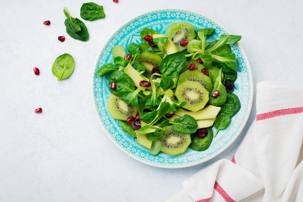 Salad of spinach baby leaves, watercress, kiwi, avocado and pomegranate in blue ceramic plate on white light wooden background. Selective focus. Top view. Copy space. - 写真・画像