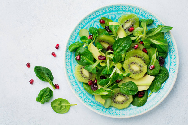 Salad of spinach baby leaves, watercress, kiwi, avocado and pomegranate in blue ceramic plate on white light wooden background. Selective focus. Top view. Copy space. - Фото, изображение