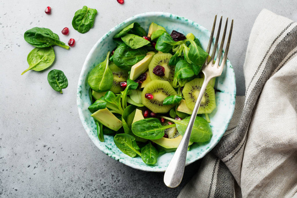 Salad of spinach baby leaves, watercress, kiwi, avocado and pomegranate in old ceramic plate on gray concrete background. Selective focus. Top view. Copy space. - Foto, imagen