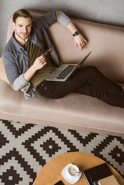 hombre guapo feliz con el ordenador portátil sentado en el sofá
 - Foto, Imagen