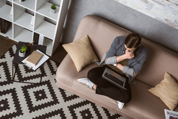 vista de ángulo alto freelancer reflexivo que trabaja con el ordenador portátil mientras está sentado en el sofá
 - Foto, imagen