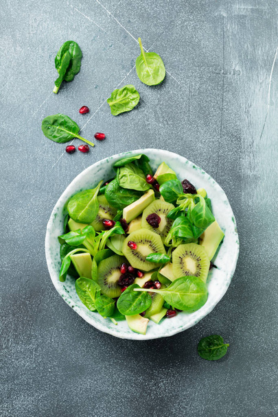 Salad of spinach baby leaves, watercress, kiwi, avocado and pomegranate in old ceramic plate on gray concrete background. Selective focus. Top view. Copy space. - Photo, Image
