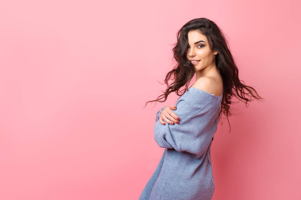Smiling woman dressed in blue knitted sweater looking over shoulder while posing on pink background - Foto, immagini