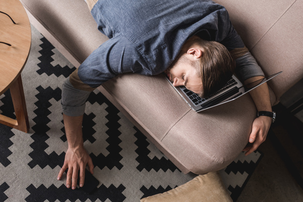 vysoký úhel pohled přepracovaný podnikatel spí na gauči s hlavou ležící na laptop - Fotografie, Obrázek