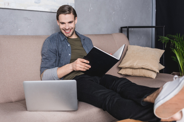 fiatal, boldog üzletember kanapén-feljegyzések olvasásához és látszó-on laptop - Fotó, kép