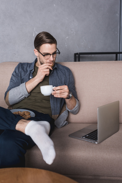 молодий красивий чоловік працює з ноутбуком на дивані під час їжі печива з кавою
 - Фото, зображення