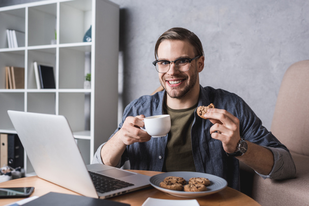 молодий красивий чоловік працює з ноутбуком і їсть печиво і каву
 - Фото, зображення