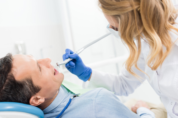 mannelijke patiënt bij tandheelkundige ingreep met behulp van tandheelkundige oefening in moderne tandheelkundige kliniek - Foto, afbeelding