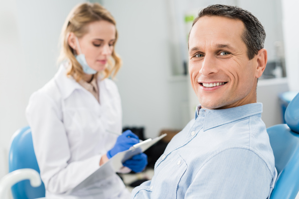 Zahnarzt nimmt Diagnose auf, während männlicher Patient in moderner Klinik wartet - Foto, Bild
