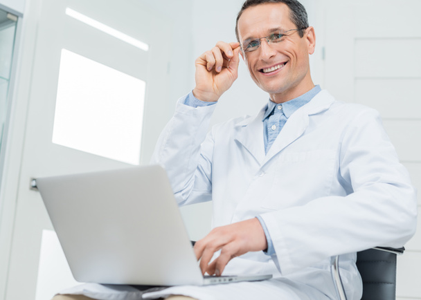 Улыбающийся врач, работающий на ноутбуке в современной клинике
 - Фото, изображение
