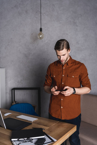 όμορφος νεαρός επιχειρηματίας χρησιμοποιώντας smartphone ενώ στέκεται κοντά στο χώρο εργασίας - Φωτογραφία, εικόνα