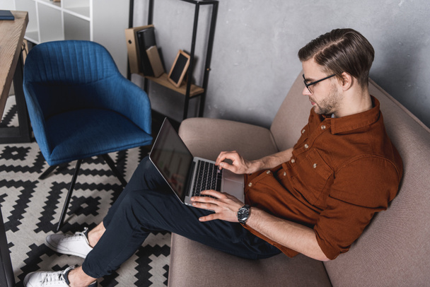 joven freelancer guapo que trabaja con el ordenador portátil en el sofá en la oficina en casa
 - Foto, imagen