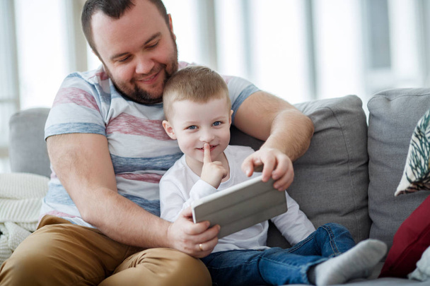 Picture of happy dad with his son with tablet sitting on gray sofa - Фото, изображение