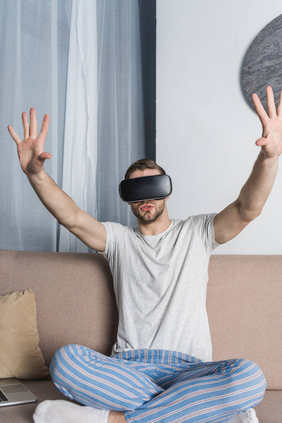 mladý muž v pyžamu a soupravu pro virtuální realitu sedí na gauči - Fotografie, Obrázek