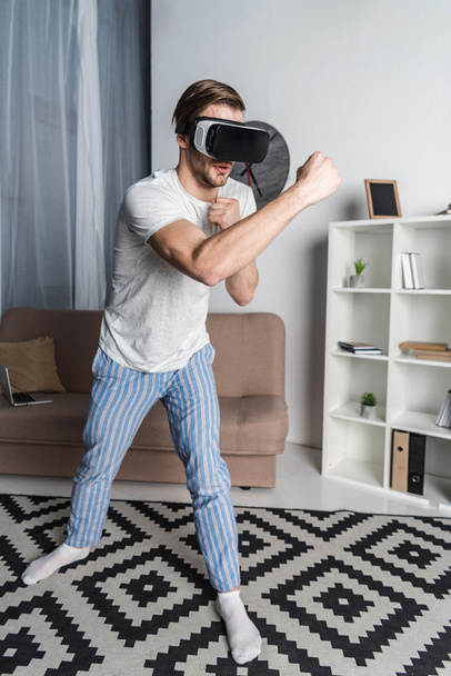 young man in virtual reality headset and pajamas playing fighting game - Фото, изображение