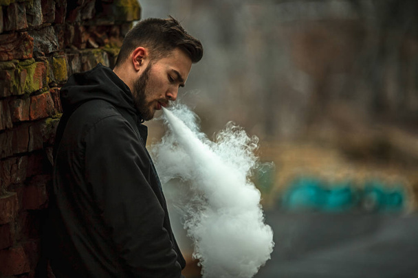 Vape man. Photo of a handsome young white guy with modern haircut in the black clothes vaping and letting off steam from an electronic cigarette near old destroyed red brick wall. Lifestyle. - Foto, Imagen