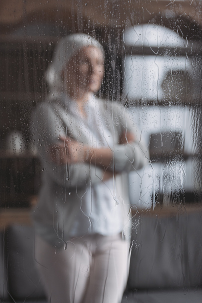 Nahaufnahme von Glas mit Regentropfen und kranker reifer Frau, die dahinter steht  - Foto, Bild