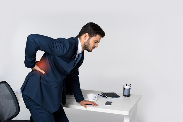 sivukuva nuoresta liikemiehestä, joka kärsii selkäkivuista ja nojaa toimistopöytään
 - Valokuva, kuva