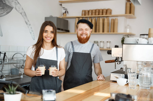 Coffee Business Concept - Positive young bearded man and beautiful attractive lady barista couple giving take away cup of coffee to custome at the modern coffee shop - Photo, Image