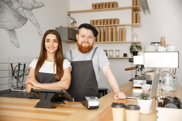 Coffee Business Concept - Positivo jovem barbudo homem e bela atraente senhora barista casal no avental olhando para a câmera enquanto está em pé no balcão do bar
. - Foto, Imagem