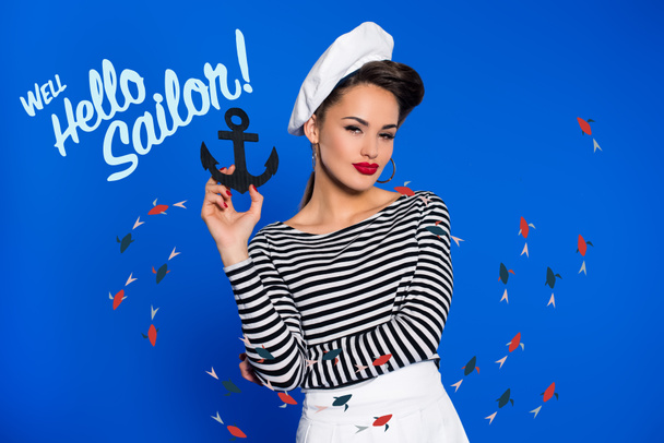 portrait of fashionable young woman in retro clothing with anchor in hand and well hello sailor inscription isolated on blue - Photo, image