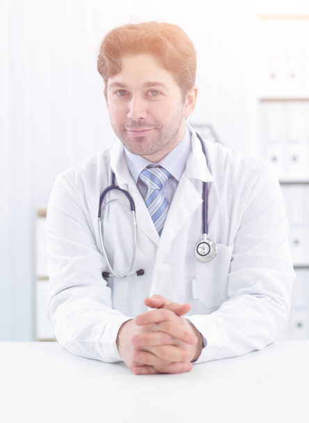 Handsome young doctor in white coat is looking at camera and smiling while standing in office - Фото, зображення