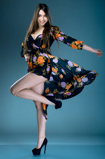 Young fashionable brunette woman in dress with floral print in high heels posing in studio. cruise collection - Fotó, kép