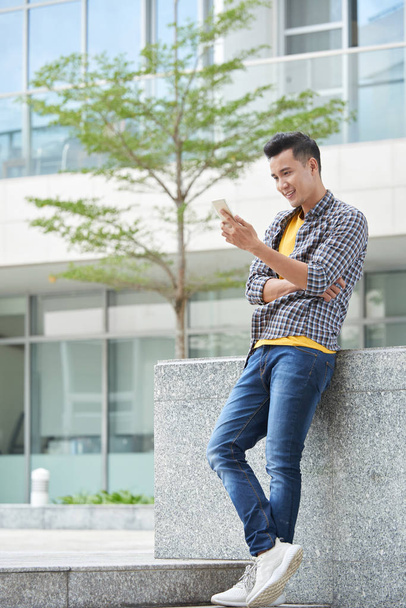 Homem vietnamita alegre em pé ao ar livre e verificando mensagens em seu telefone
 - Foto, Imagem