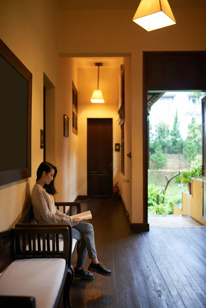 Nuori aasialainen nainen istuu penkillä hotellihuoneessa ja lukee kirjaa
 - Valokuva, kuva