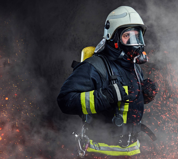 Retrato de bombero vestido de uniforme y una máscara de oxígeno de pie en chispas de fuego y humo sobre un fondo oscuro
. - Foto, imagen