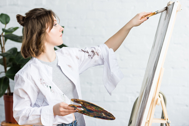 Young creative girl smears paint on canvas in light studio - Photo, Image