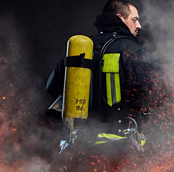 Un pompier avec un réservoir d'oxygène sur le dos debout dans des étincelles de feu et de la fumée sur un fond sombre
. - Photo, image