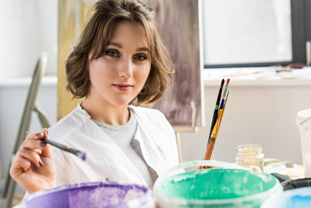 Young artistic girl dunks a paintbrush into the paint in light studio - 写真・画像