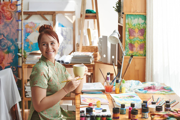 Gelukkig jonge vrouw drinken thee en schilderen in haar atelier - Foto, afbeelding