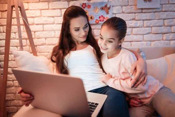 Mother and little daughter watching movies on laptop under lampshade at night at home. - Photo, image