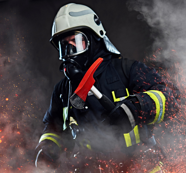 Un bombero vestido con un uniforme y una máscara de oxígeno sostiene un hacha roja de pie en chispas de fuego y humo sobre un fondo oscuro
. - Foto, Imagen