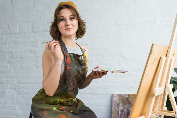 Молодая вдохновленная девушка, работающая с ножом за мольбертом в легкой студии
 - Фото, изображение
