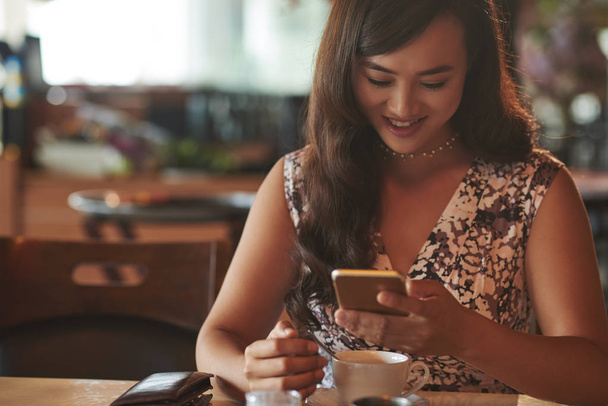 Улыбаясь, красивая азиатка читает новости на смартфоне, когда пьет кофе.
 - Фото, изображение