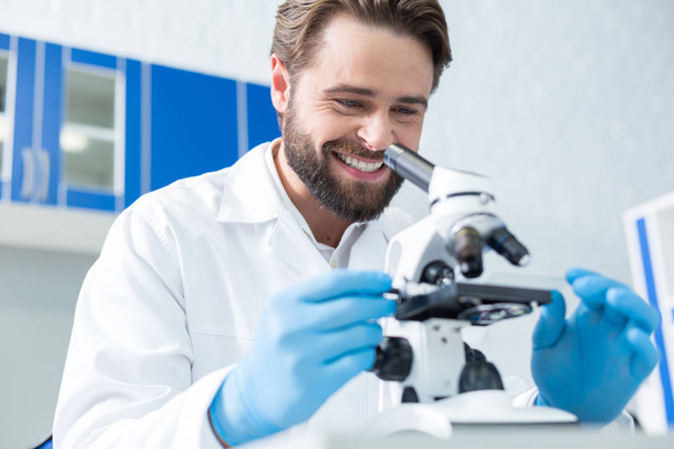Professional biologist. Happy cheerful positive scientist working with the microscope and smiling while enjoying his job - 写真・画像