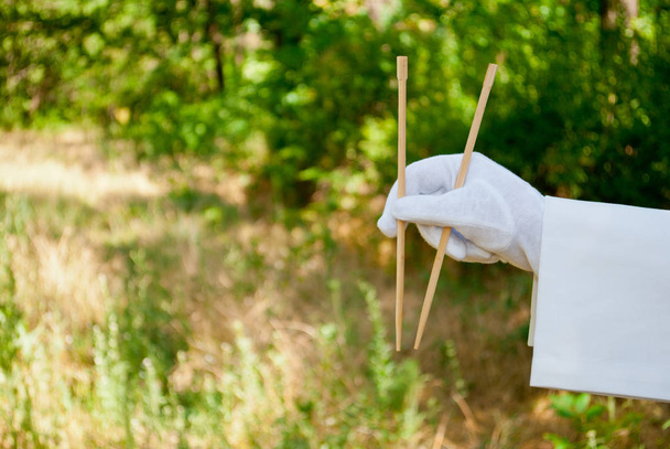 The hand of the waiter in a white glove and with a white napkin holds the narrow long wooden sticks for the oriental cuisine on a blurred background of nature green bushes and trees - Foto, Bild