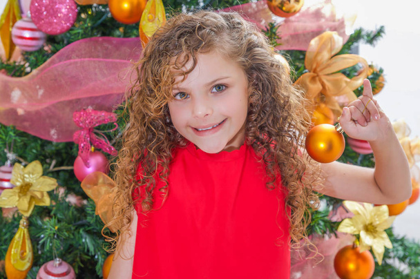 Close up of happy girl, wearing a red blouse and holding a christmas ball in front of a Christmas tree, christmas concept - Фото, изображение