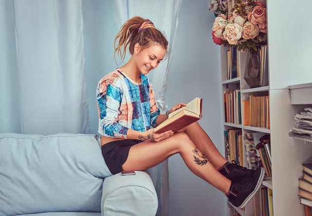 Young tattooed girl read a book sitting on a sofa at home. - Foto, imagen